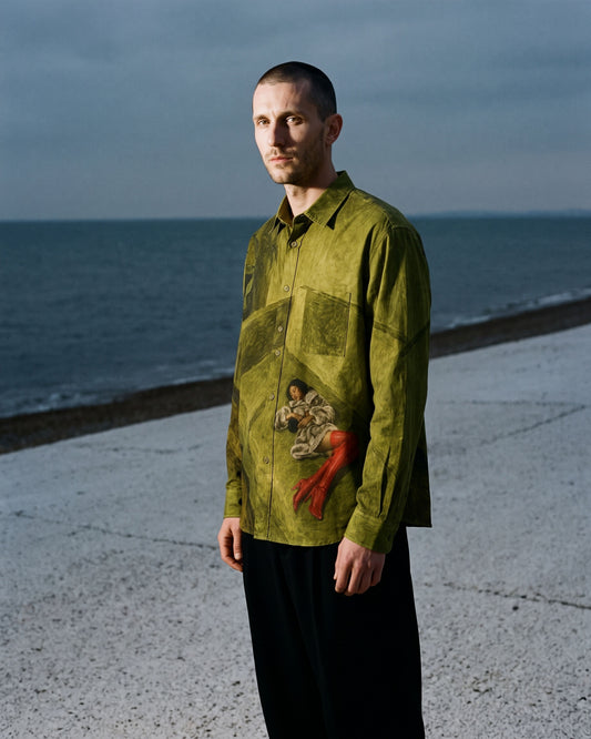 A man stands on a concrete pier overlooking the ocean, wearing a long-sleeve button-up shirt in olive green with a vintage-style painting print on the front pocket and black trousers.
