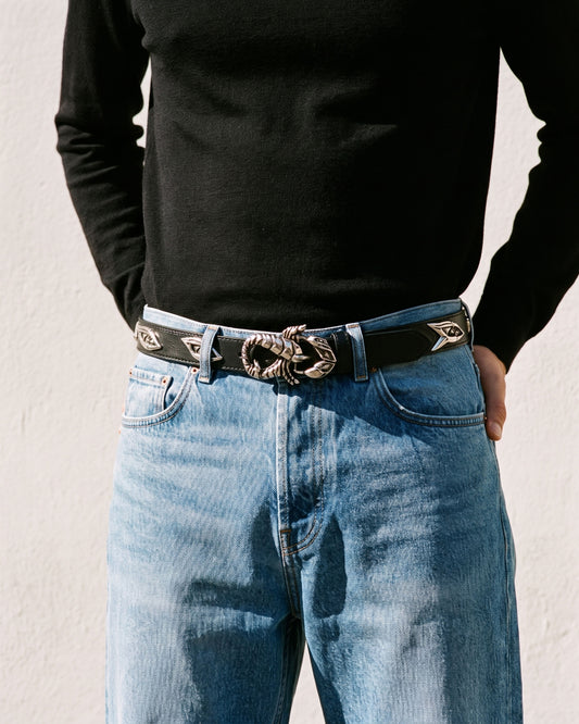 A close-up of a person wearing a black long-sleeve shirt, light blue jeans, and a black leather belt with a large silver snake buckle and silver star-shaped belt tips, standing against a white wall.