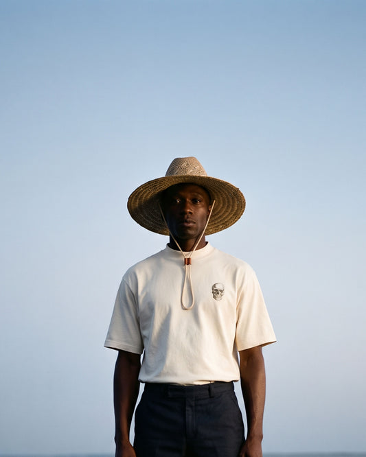 A man stands barefoot on a pier by the water, wearing a wide-brim straw hat, a cream short-sleeve polo shirt with a drawstring neck, and dark shorts against a calm blue sky and sea background.