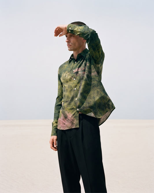 A man stands in a minimalist desert landscape wearing a long-sleeve button-up shirt with a green and pink abstract print, black trousers, and a matching patterned headband.