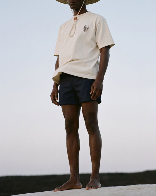 A model wearing a cream-colored short-sleeve t-shirt with a small chest graphic, navy blue shorts, and a wide-brimmed straw hat, standing outdoors against a pale sky.
