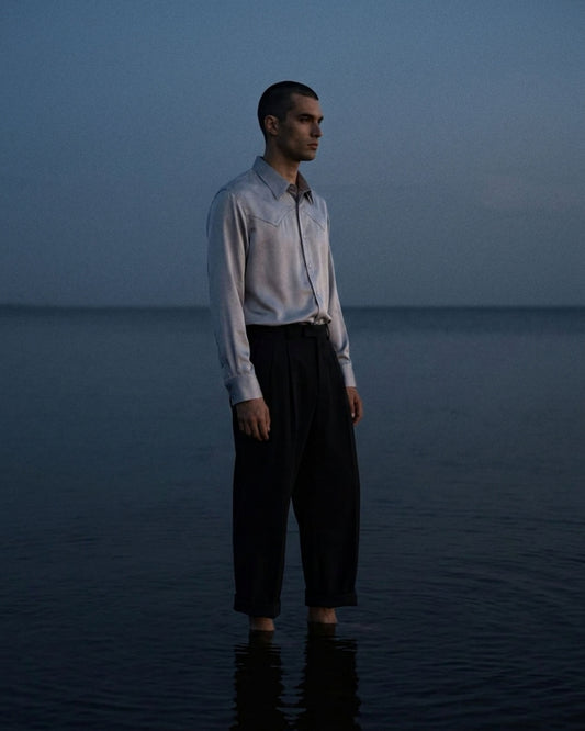 A man stands barefoot in shallow water at dusk, wearing a light-colored long-sleeve button-up shirt, dark trousers, and a tie.